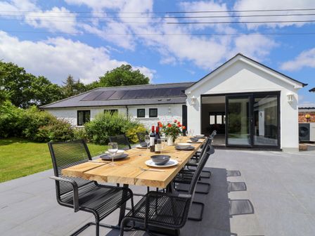 An outdoor patio dining area with a wooden table set with plates glasses and bottles in front of a white house with black window frames and solar panels on the roof at Hartgrove Farm Bungalows Musbury