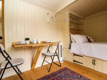 A small room with a wooden table set for two with chairs beside a bed with white linens and built-in shelves at Mitford Gypsy Hut in Moreton-In-Marsh