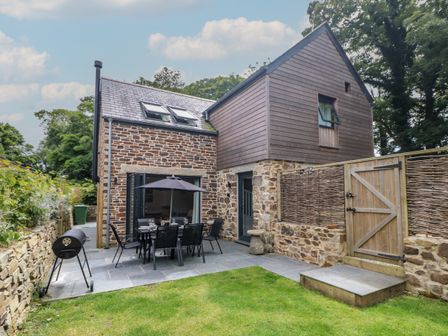 An outdoor patio area with a dining table and umbrella a barbecue grill and a stone and wooden house in the background at Wildwood in Newquay