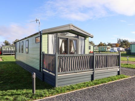A green mobile home with a small porch and glass doors on a grassy plot with other similar units nearby