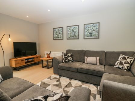 A living room with a grey sectional sofa a patterned rug a wooden TV stand with a television and three paintings of trees on the wall at Brimstone in Backbarrow near Ulverston