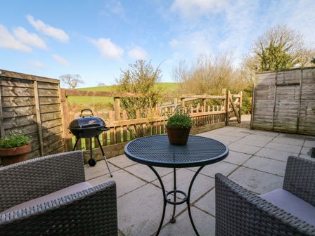 A patio with two wicker chairs a round metal table with a plant a barbecue grill and wooden fences at The Linhay in Muddiford near West Down
