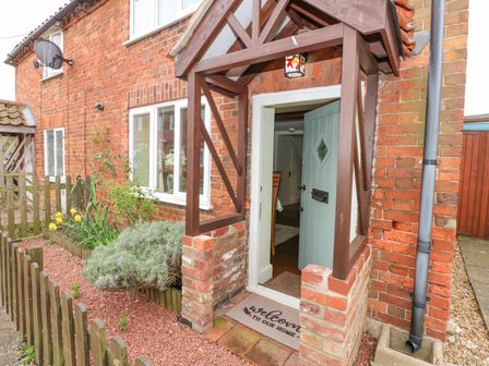 A brick house entrance with a wooden porch and a blue door partially open at Easter Cottage in Dersingham