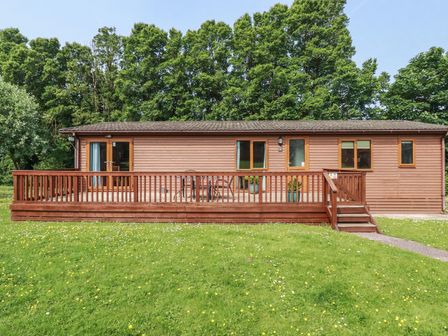 A wooden cabin with a deck and stairs in a grassy area with trees in the background at 4 The Glade in St Minver