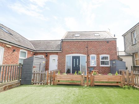 A brick house with two black doors two windows two garden sheds wooden fences and planter boxes on a lawn at Pine Cottage in Dinas Dinlle