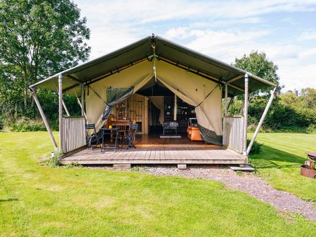 A tent with an open front showing chairs a table a wood stove and wooden shelves at Beech in Dalwood