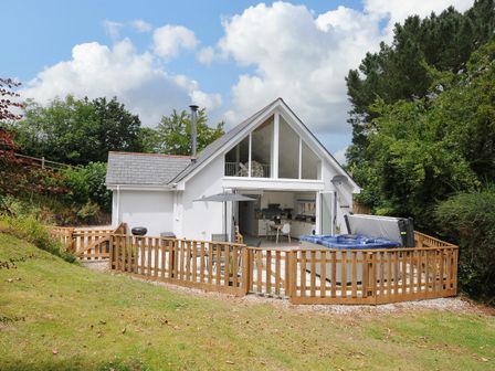 A white house with large windows a fenced patio with a hot tub and outdoor furniture surrounded by trees at Otters Leap in Bideford