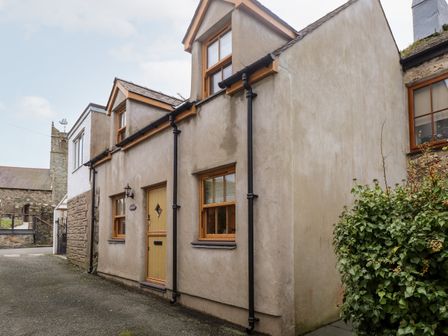 The exterior of a two-story house with beige walls wooden framed windows and a yellow door by a narrow street with a stone church in the background at Bull Bach Old Smithy in Nefyn