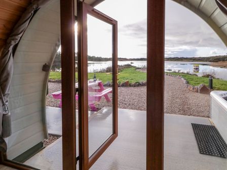 An open wooden door leading to an outdoor gravel area with a pink picnic table and a bottle by a lake at The Wheelhouse Pod No 2 in Burtonport County Donegal