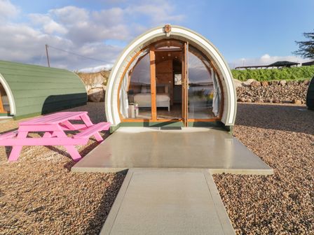 A small curved cabin with glass doors opening to a bed inside a gravel area with a pink picnic table at The Wheelhouse Pod No 7 in Burtonport County Donegal