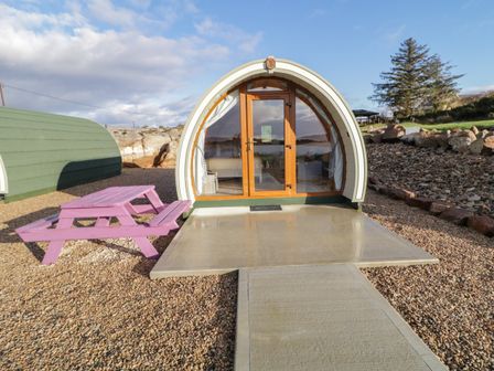 A curved pod structure with glass door and windows on a concrete patio with a purple picnic table on gravel at The Wheelhouse Pod No 9 in Burtonport County Donegal