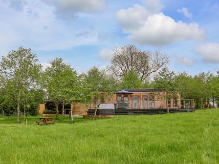 A wooden house with a deck, an umbrella, a hot tub, trees, and a picnic bench on grass at Holmside in Audley Brow near Market Drayton