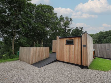 A wooden cabin with a gravel driveway and trees in the background at Herdwick Cabin in Hutton John near Penruddock
