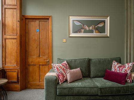 A green sofa with patterned and plain cushions next to a wooden door with a painting of two rabbits on the wall above