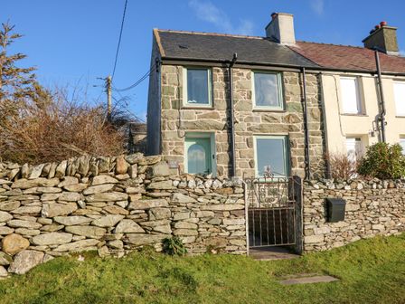 A stone house with green window frames and door behind a stone wall with a metal gate at Fron Fair in Penygroes