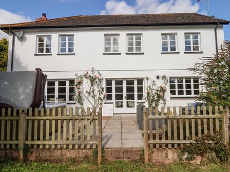 A white two-story house with multiple windows a wooden fence and a paved patio with climbing plants at Culver Court Cottage in Venny Tedburn near Crediton
