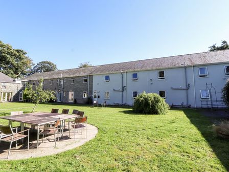 An outdoor garden area with a large green lawn surrounded by a two-story building with multiple windows and doors and a wooden patio table with chairs at The Bevan in Bontnewydd Gwynedd
