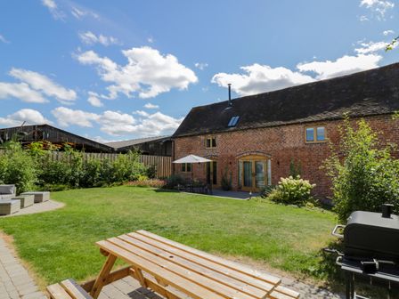 A garden with a wooden picnic table a barbecue and a patio set with an umbrella outside a brick building at The Stables in Droitwich Spa