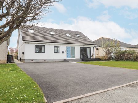 A detached house with a dark tiled roof and white walls with a light blue front door and large driveway with lawn on both sides at Blue Bay Villa in Trearddur Bay