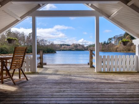 A wooden deck with railing and a chair overlooking a lake with trees in the background at Lilymere Boat House in Sedbergh