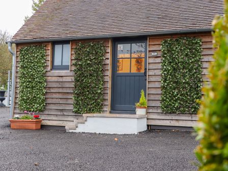 The exterior of a wooden cottage with a blue door steps and trellises with green plants at Bee Cottage in Ottinge near Lyminge