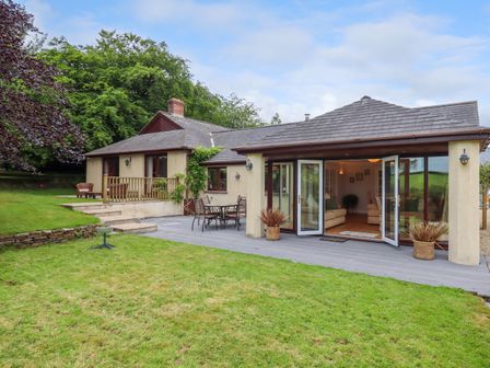 A house with patio doors open to a living room and an outdoor table with chairs on a deck surrounded by grass and trees at Hole Park in Holsworthy