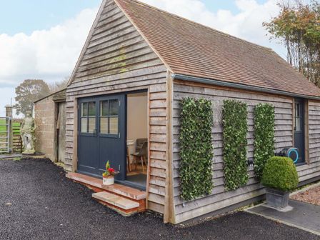 A small wooden building with a double door open showing a table and chairs inside and plants on the exterior walls at Kingfisher Cottage in Ottinge near Lyminge
