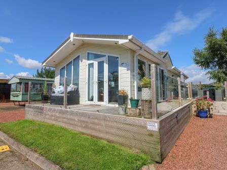 A small house with a wooden deck and glass railing surrounded by grass and gravel at A and C Lodge-Plot 21 in Armathwaite