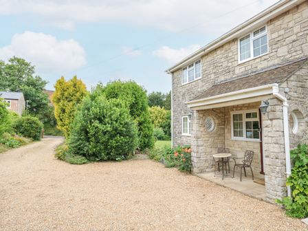 A stone house with a small porch table and chairs next to a gravel driveway surrounded by bushes at Glyde House in Dorchester
