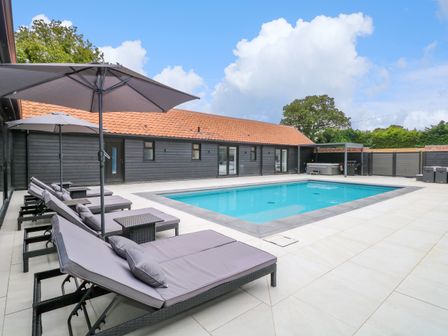 An outdoor swimming pool with lounge chairs and umbrellas next to a black building at County Farm Barn in South Walsham
