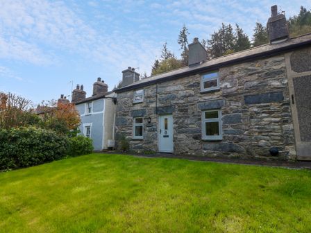 A stone cottage with a white door and front lawn surrounded by trees and bushes at 5 Banc Llugwy in Betws-Y-Coed