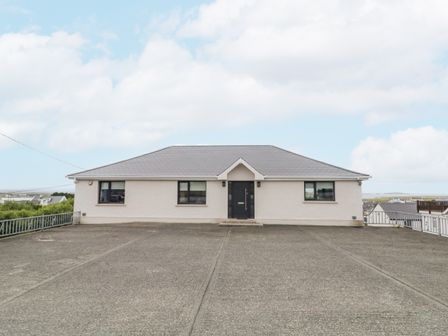 A single-story house with a dark front door and three windows facing a large concrete driveway at Cottage Catriona in Derrybeg County Donegal