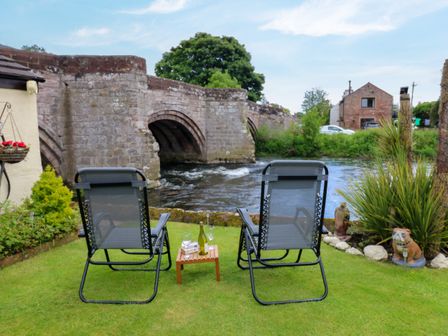 Two chairs and a small table on grass facing a stone bridge over a river with a house in the background at River Cottage in Eamont Bridge