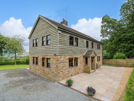 An exterior view of a two-story house with stone and wood siding a paved patio and gravel driveway at Oak Apple Cottage in Presteigne