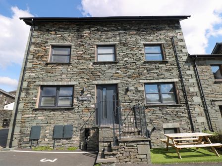 The exterior of a stone building with six windows a black door a wooden picnic table and a parking area at Quarters in Ambleside