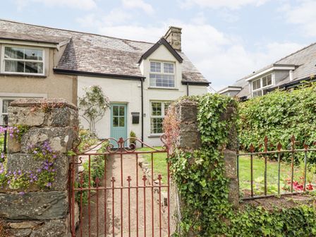 Front yard with a pathway, a gate, stone pillars with plants, and a two-story house with a pale blue door at Ivy Cottage in Harlech