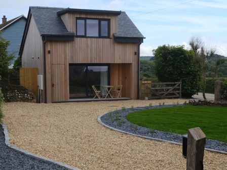 A wooden house with a gravel driveway two chairs and a table a green lawn and a wooden gate at The Cabin at Ropewalk in Goonbell near St Agnes