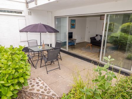 An outdoor patio area with a table, chairs, and umbrella next to a sliding glass door leading to a living room with a sofa, TV, and wooden floor at 14 Coedrath Park in Saundersfoot