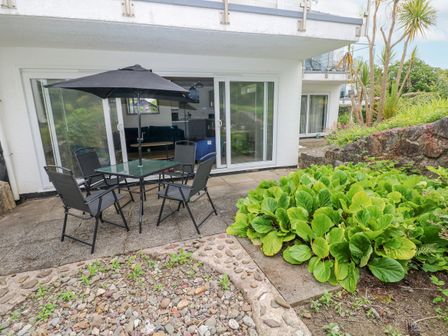 A patio area with a glass table and four chairs under a black umbrella next to green plants and sliding glass doors at 29 Coedrath Park in Saundersfoot