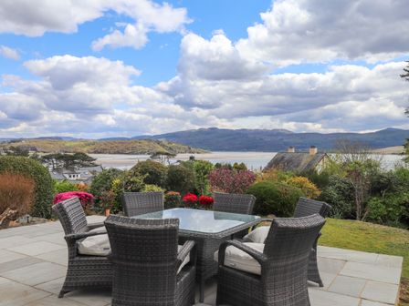 An outdoor patio with a glass table and six wicker chairs overlooking a garden and a body of water with hills in the background at Bro Gwylan in Borth-y-Gest near Porthmadog