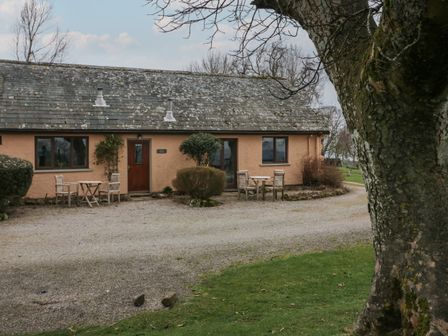 A single-story cottage with two outdoor seating areas on a gravel driveway and a large tree in front at Little Winder in Askham Cumbria