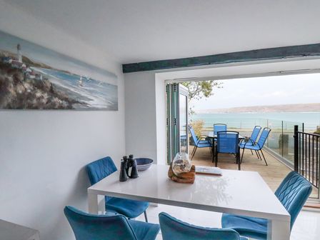 A dining area with a white table and blue chairs next to an open patio door overlooking a deck with a table and blue chairs and sea view at The Cubby Hole in Sennen Cove