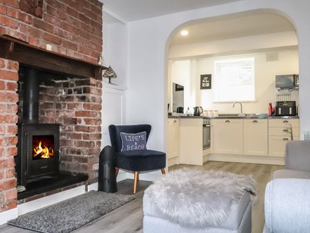 A living room with a brick fireplace and a navy chair with a pillow and an open kitchen in the background at Henllys in Harlech