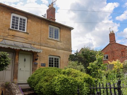 A stone cottage with green door and windows surrounded by bushes and garden at Blossom Cottage in Chipping Campden