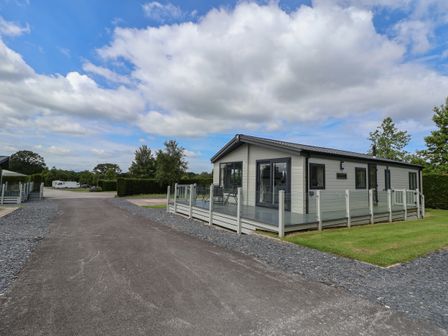 A single-story prefabricated house with a glass railing and a patio on a grassy plot by a paved road at Lodge 3 in St Asaph