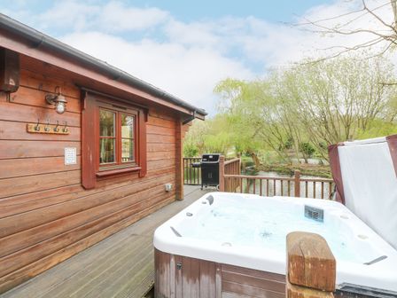 An outdoor wooden deck with a hot tub and a barbecue grill next to a wooden cabin by a body of water at Kingfisher Lodge in Badwell Ash