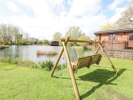 A wooden swing overlooking a pond with a wooden cabin nearby at Wagtail Lodge in Badwell Ash