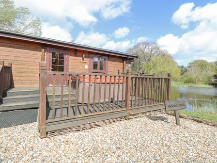 A wooden cabin with a fenced deck and a hot tub near a pond with trees at Woodpecker Lodge in Badwell Ash