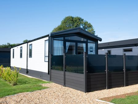 A modern static caravan with black trim and a glass panel fence on a gravel pathway surrounded by grass at Tiggywinkles in Chipping Norton