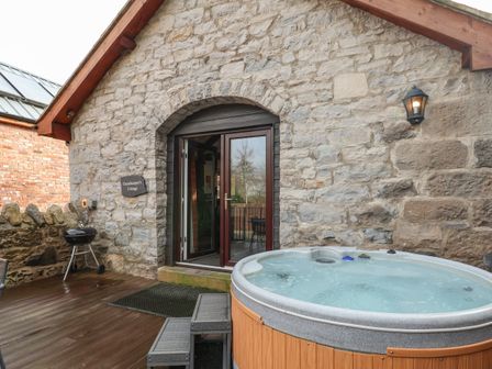 An outdoor area with a stone wall building a round hot tub and a barbecue grill at Gamekeepers Cottage in Llys Llannerch St Asaph